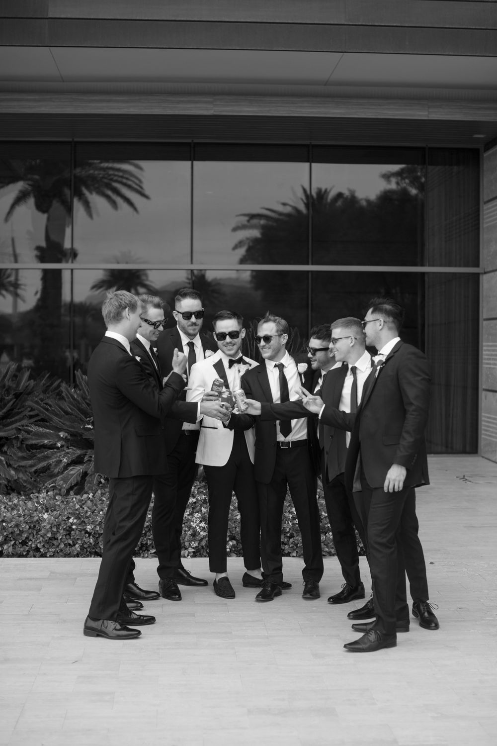 Groomsmen in black suits gather outside a modern building for casual group photos.