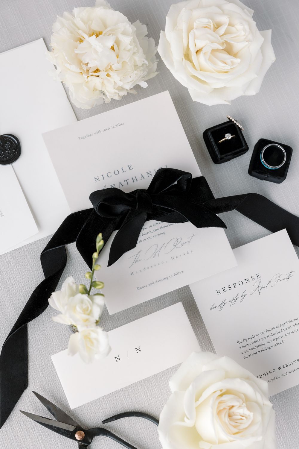 Elegant black and white wedding invitation suite with velvet ribbon, white roses, and ring boxes on gray background.