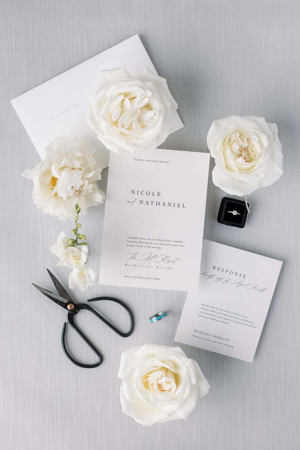 Modern minimalist wedding invitation suite for Nicole and Nathaniel with white roses, scissors, and ring box on gray linen.
