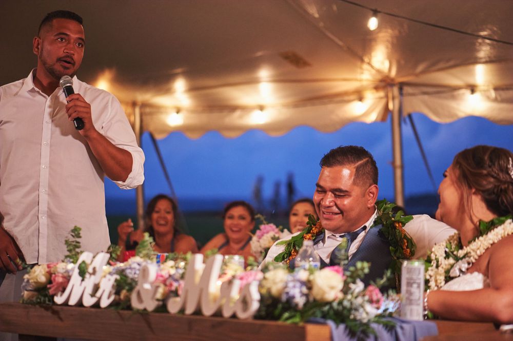 Wedding guests enjoy speeches and toasts during an outdoor evening reception under string lights.