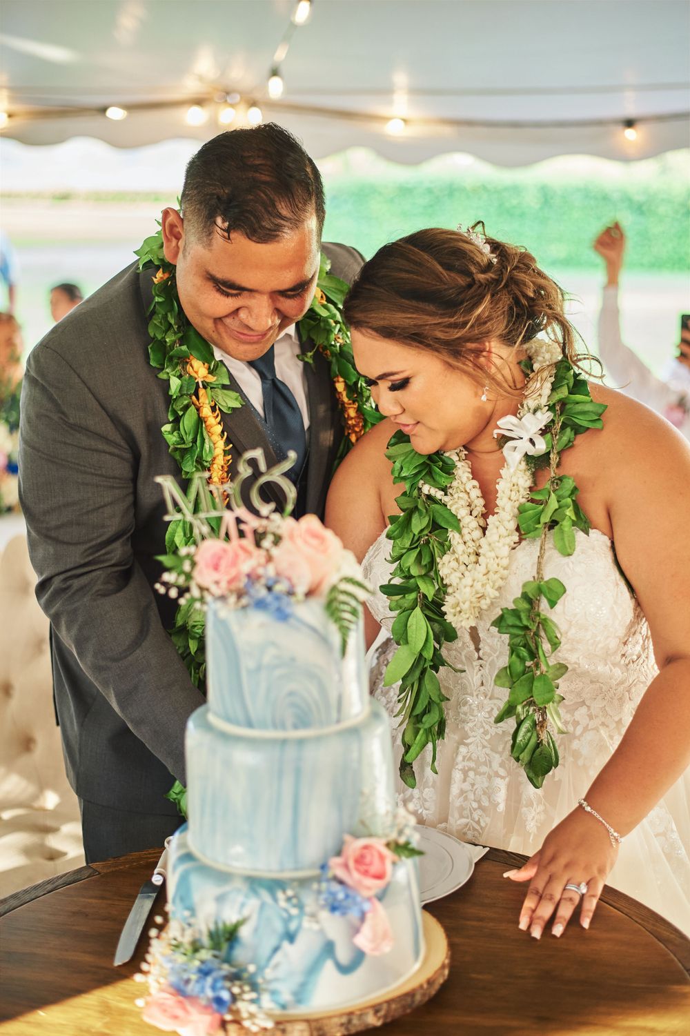 A couple wearing green leis cuts into their blue and white wedding cake together.