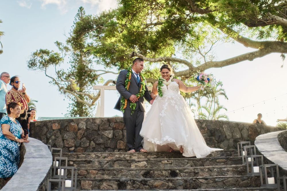 A wedding couple descends stone steps at an outdoor venue under a large tree.
