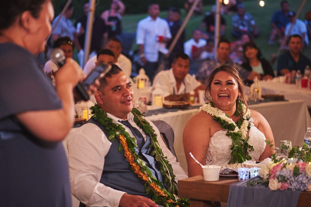 A couple wearing traditional Hawaiian leis shares a joyful moment during their wedding reception under a tent.