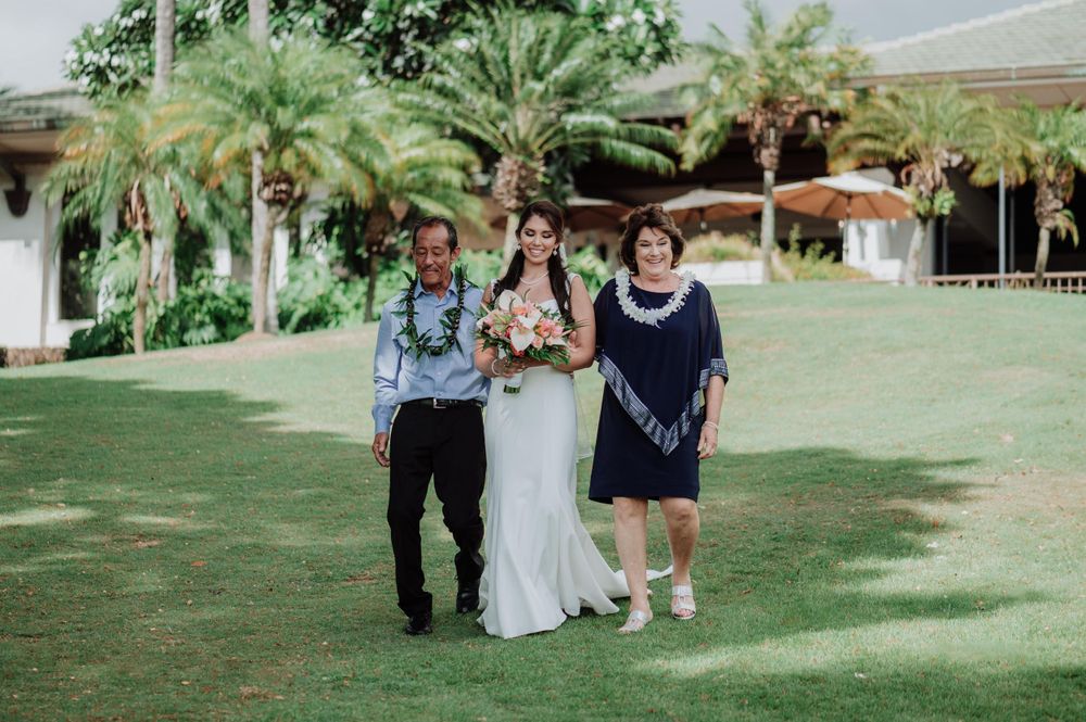 Wedding ceremony procession on grassy lawn with white arbor and palm trees.