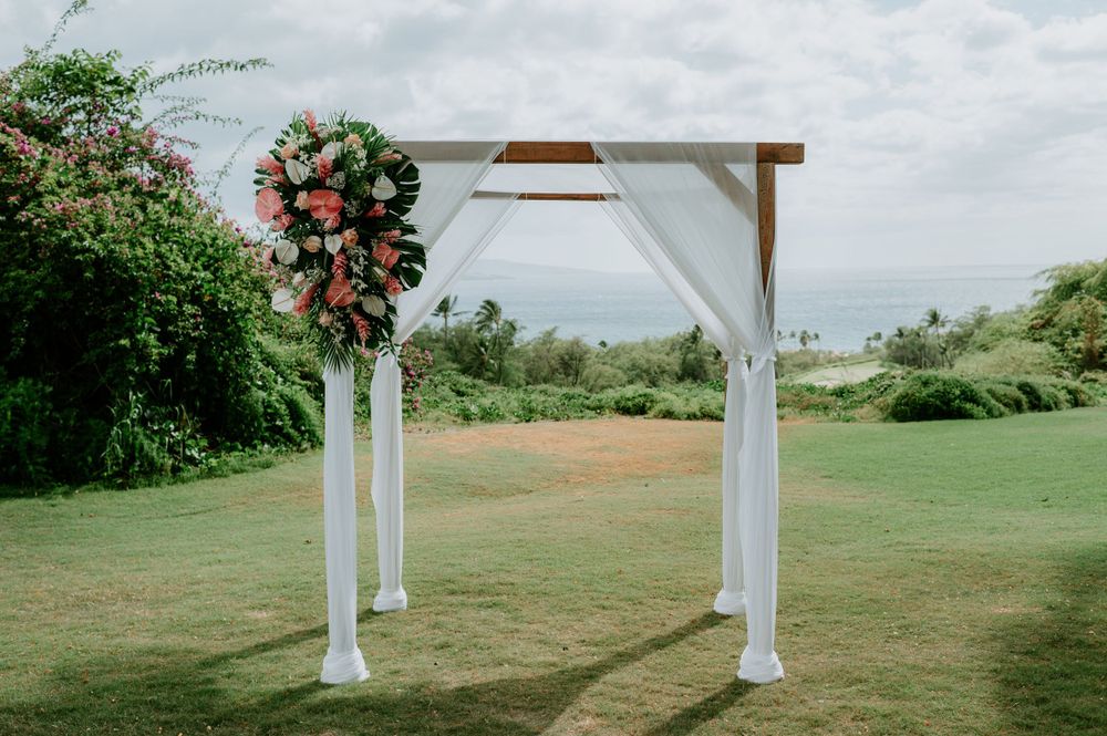 White wedding arbor decorated with pink flowers stands on a grassy lawn under cloudy skies.