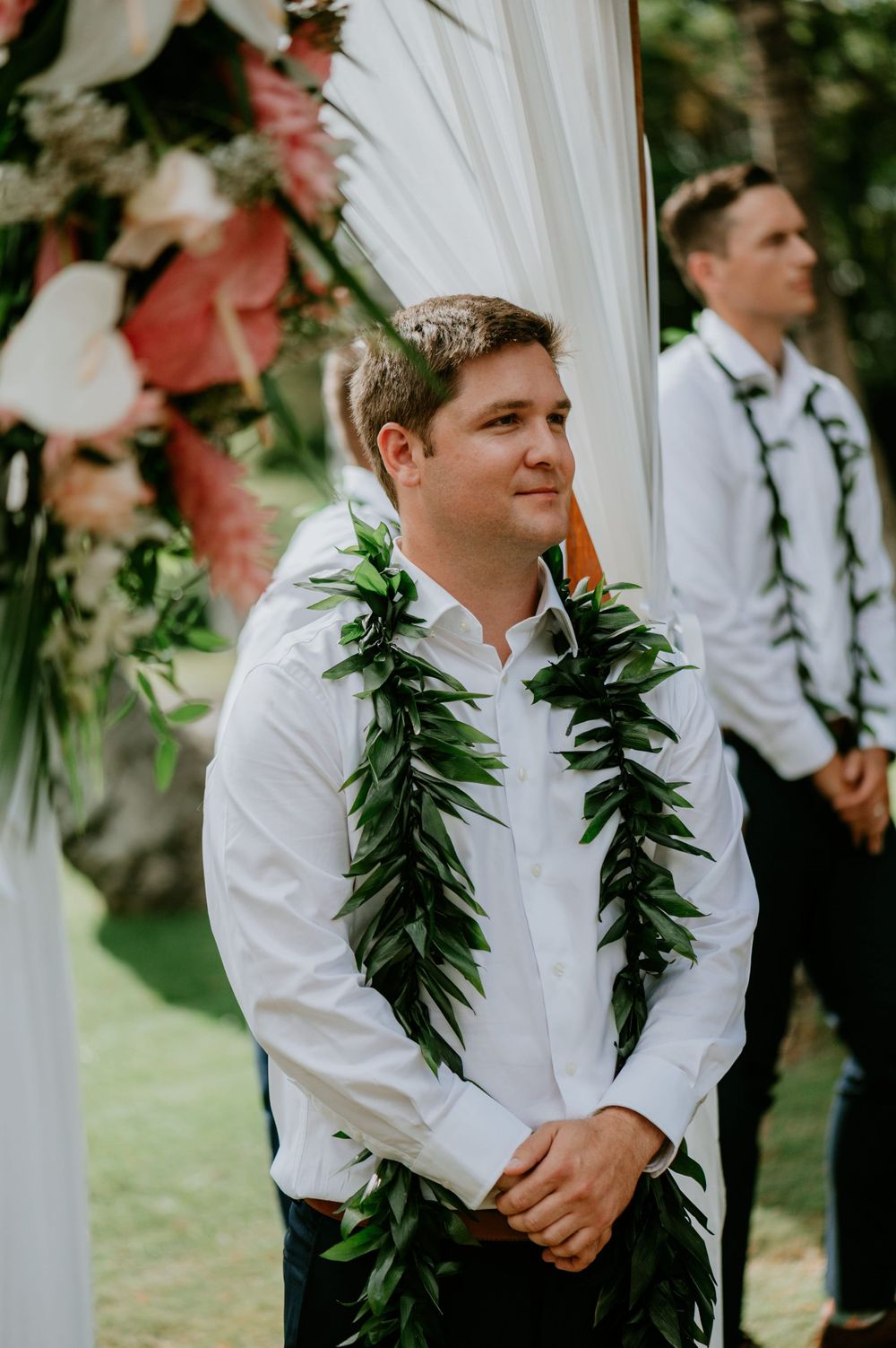 Groomsmen wearing white shirts and green leaf leis stand at outdoor wedding ceremony.