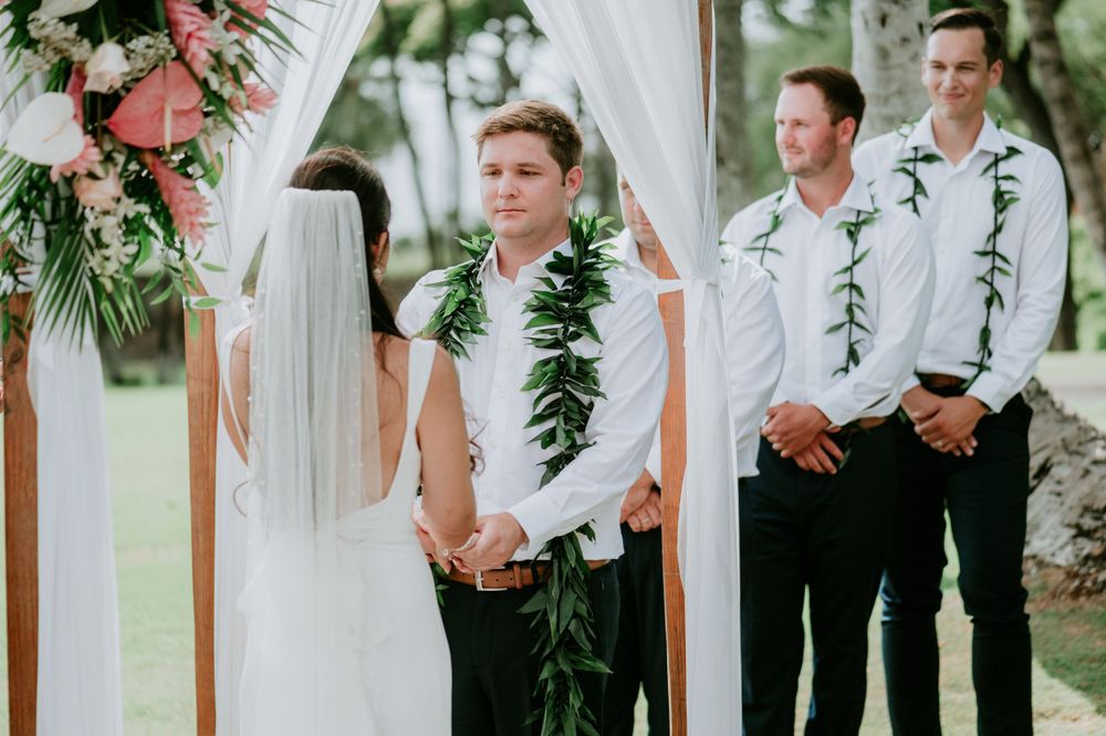 Wedding party members stand under a white draped arch decorated with tropical greenery.