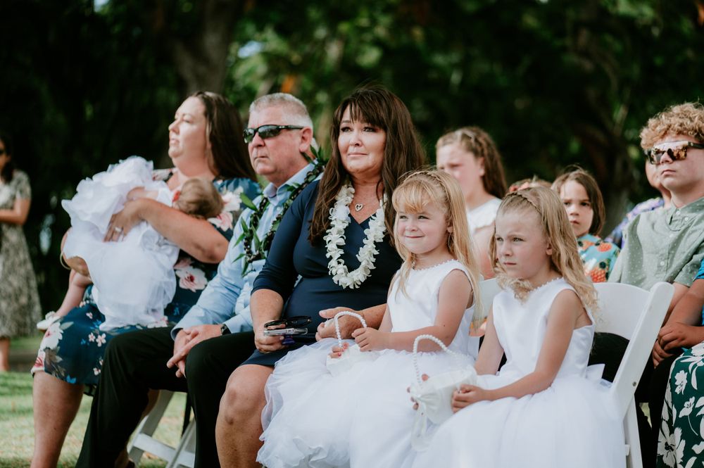 Wedding guests seated in white chairs watch an outdoor ceremony under palm trees.