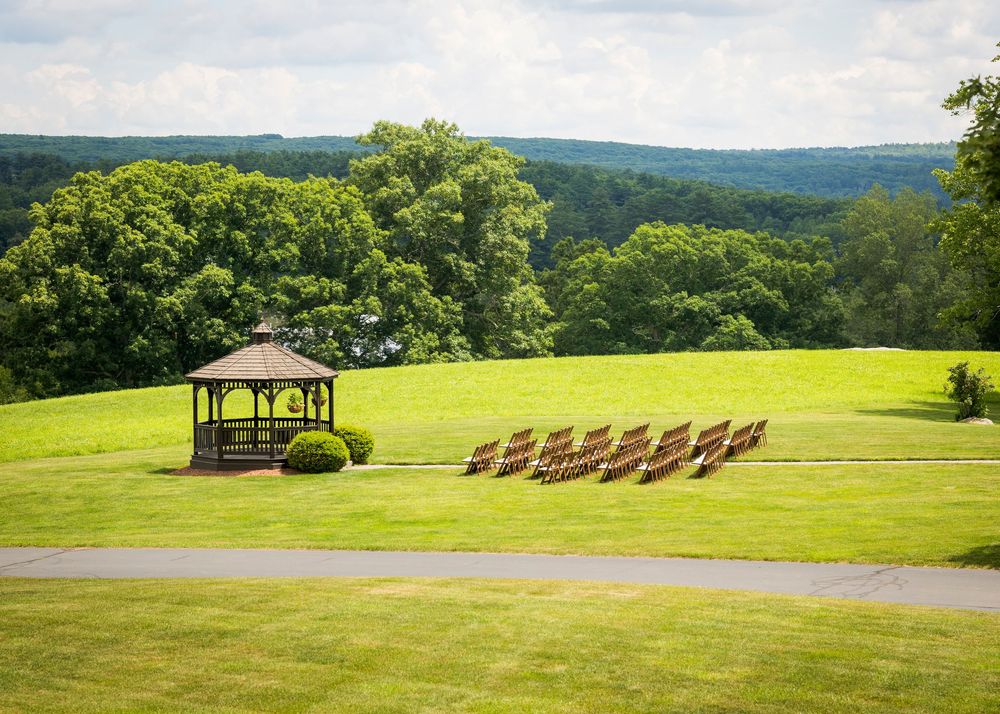 Outdoor wedding ceremony setup with wooden chairs facing a gazebo on a lush green hillside with forest views.