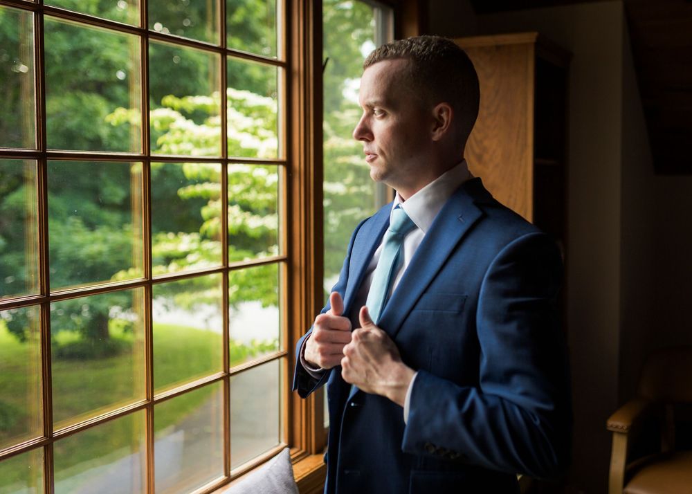 Groom in navy suit adjusts tie while gazing out window before wedding.