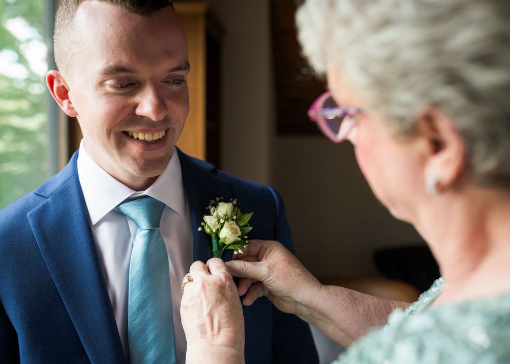 Smiling groom in navy suit gets boutonniere pinned by older woman before wedding ceremony.
