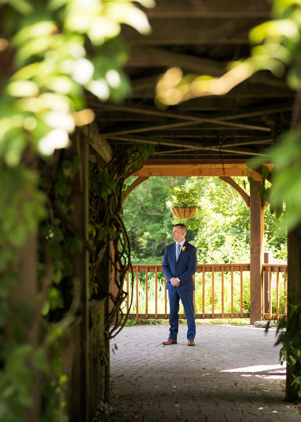Groom in navy suit standing under rustic wooden pergola surrounded by lush green foliage at outdoor wedding venue.