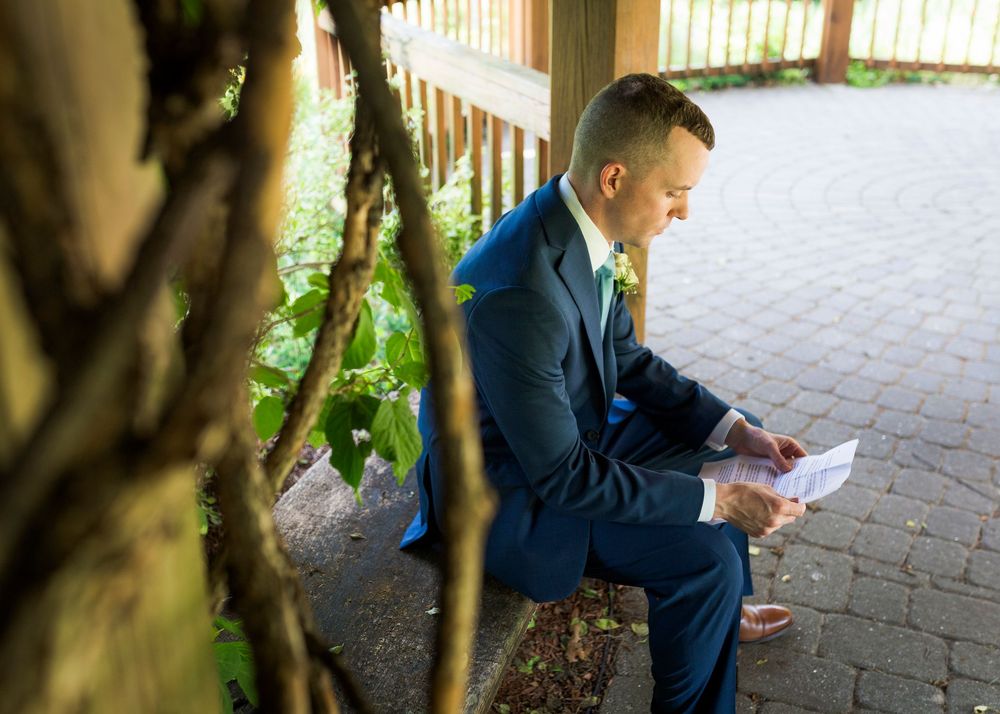 Groom in navy suit sitting outdoors reading a note before his wedding ceremony in a garden setting.