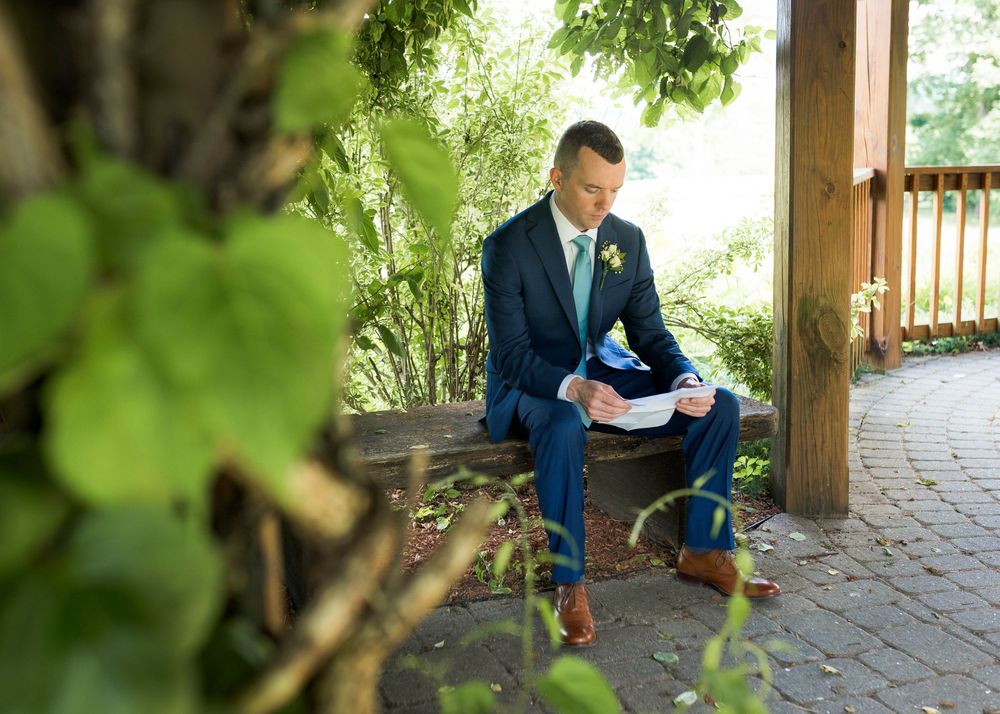 Groom in navy suit reads vows on bench surrounded by lush greenery.