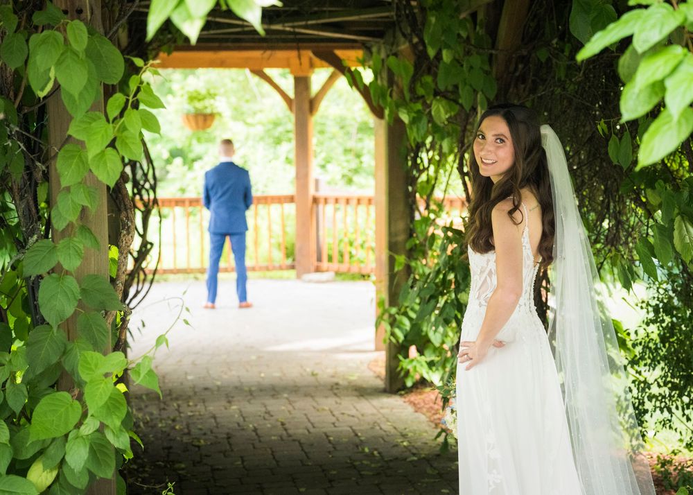 Bride in white gown and veil stands under ivy-covered arbor as groom in blue suit waits ahead.