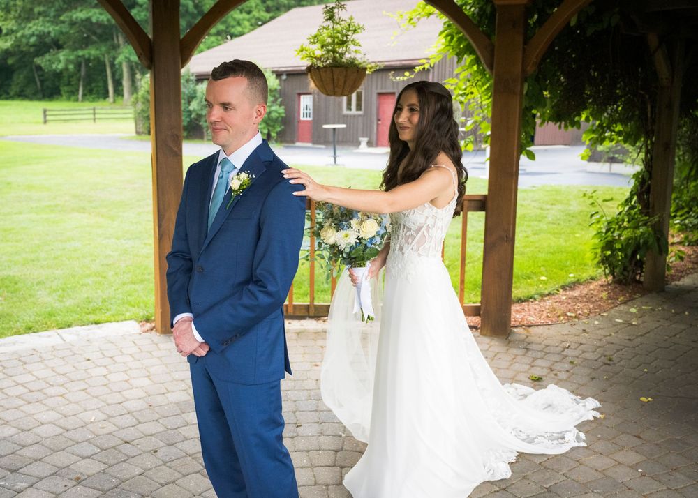 Bride in white gown taps groom's shoulder during first look under wooden gazebo at outdoor wedding.