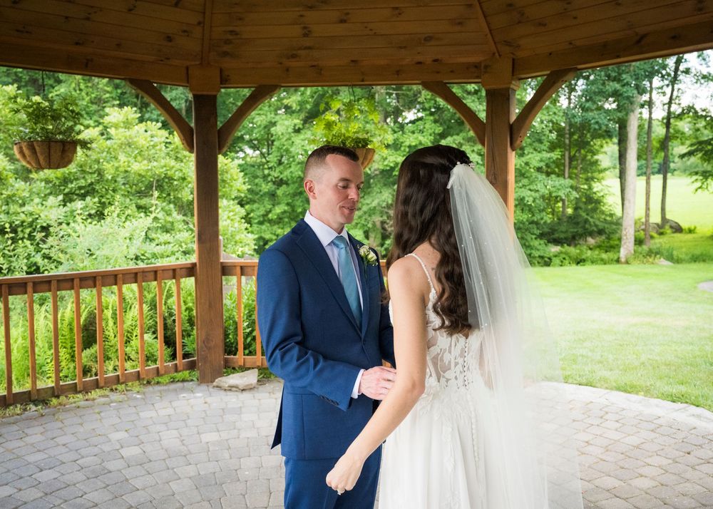 Groom in blue suit and bride in white veil share first look moment under rustic gazebo surrounded by greenery.