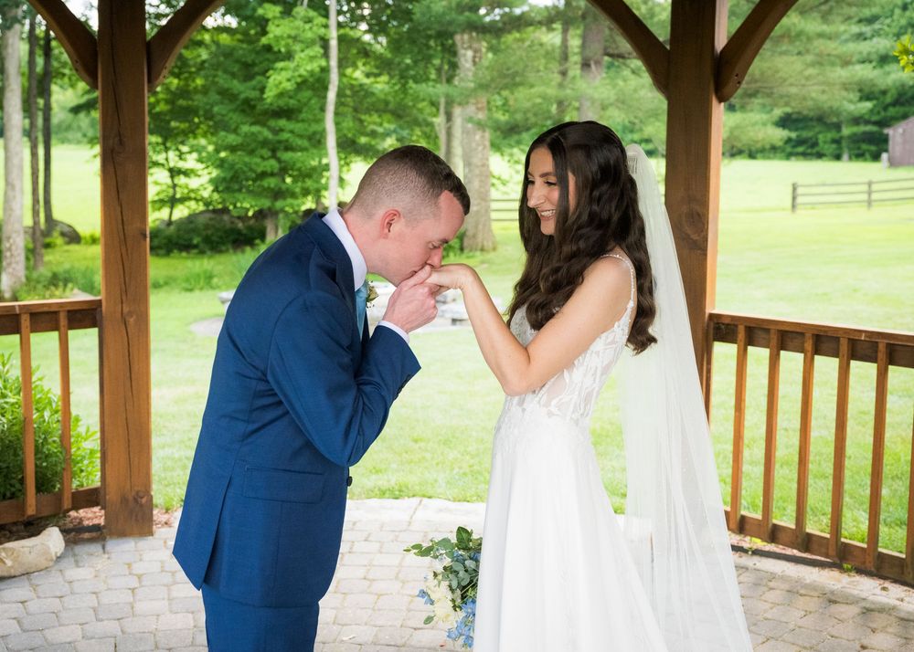 Groom kissing bride's hand under wooden gazebo during outdoor wedding ceremony.