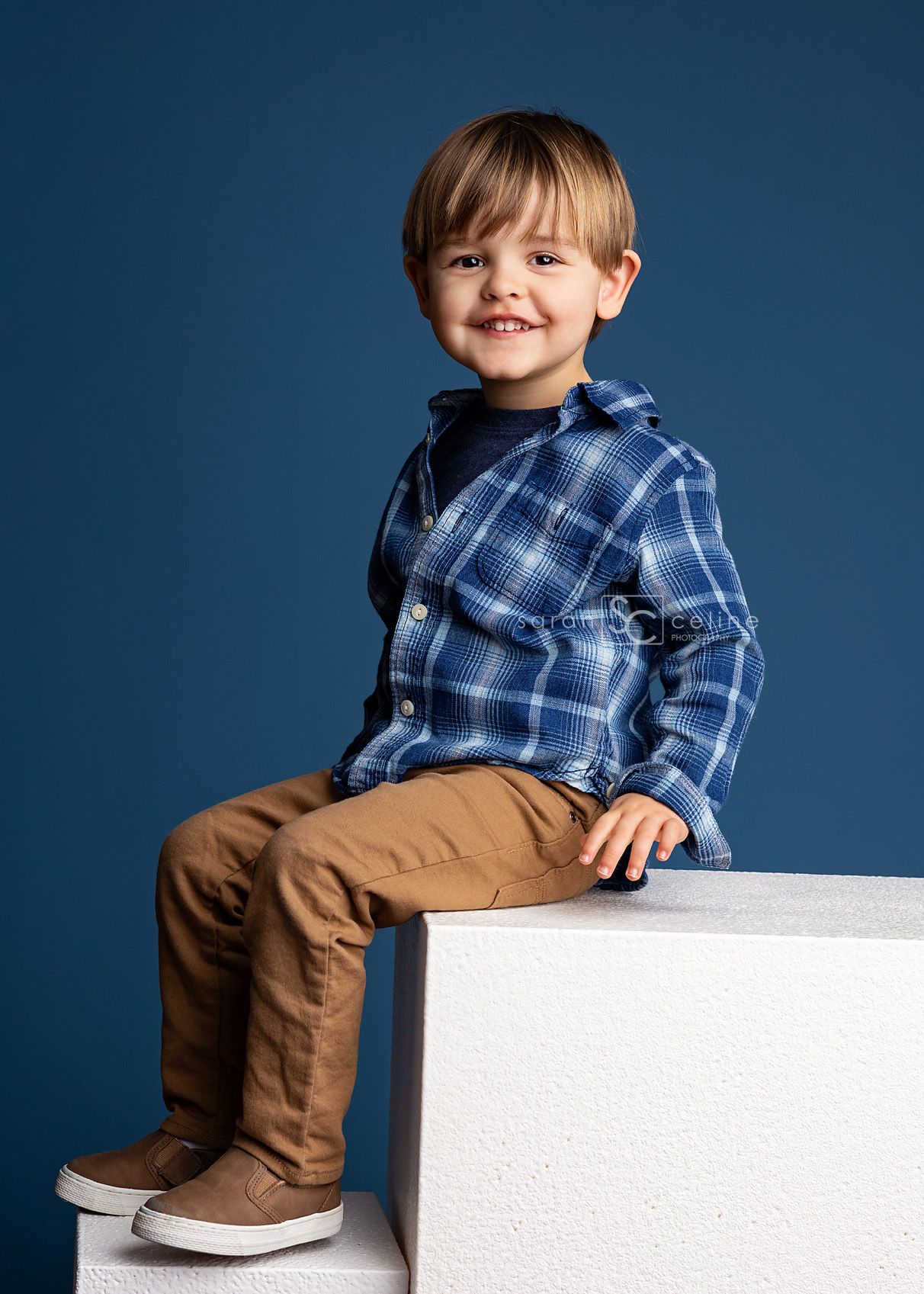 Child wearing a blue plaid shirt and brown pants sits casually on a white platform against a dark blue backdrop.