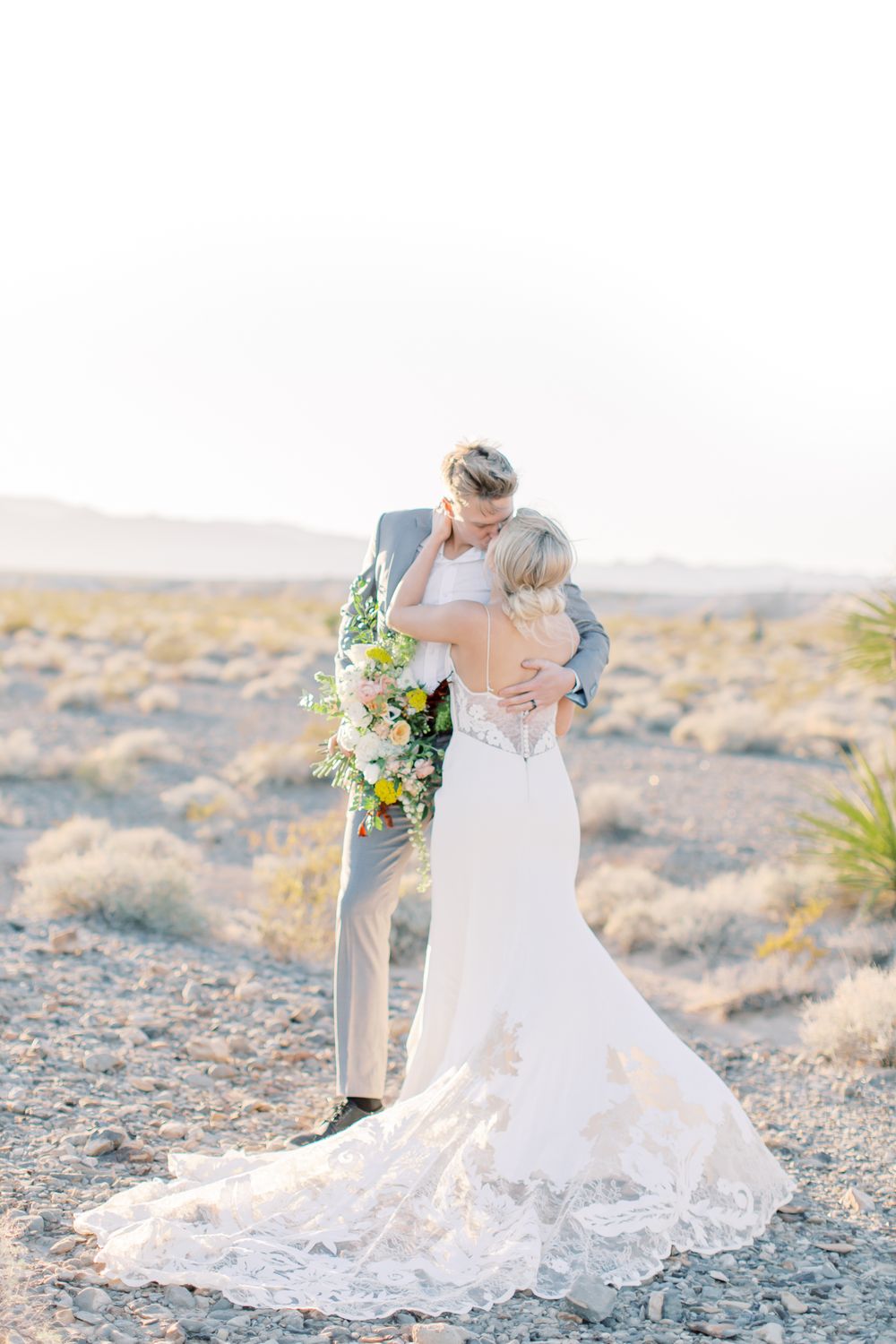 A romantic desert wedding portrait featuring a flowing lace train and elegant floral bouquet.