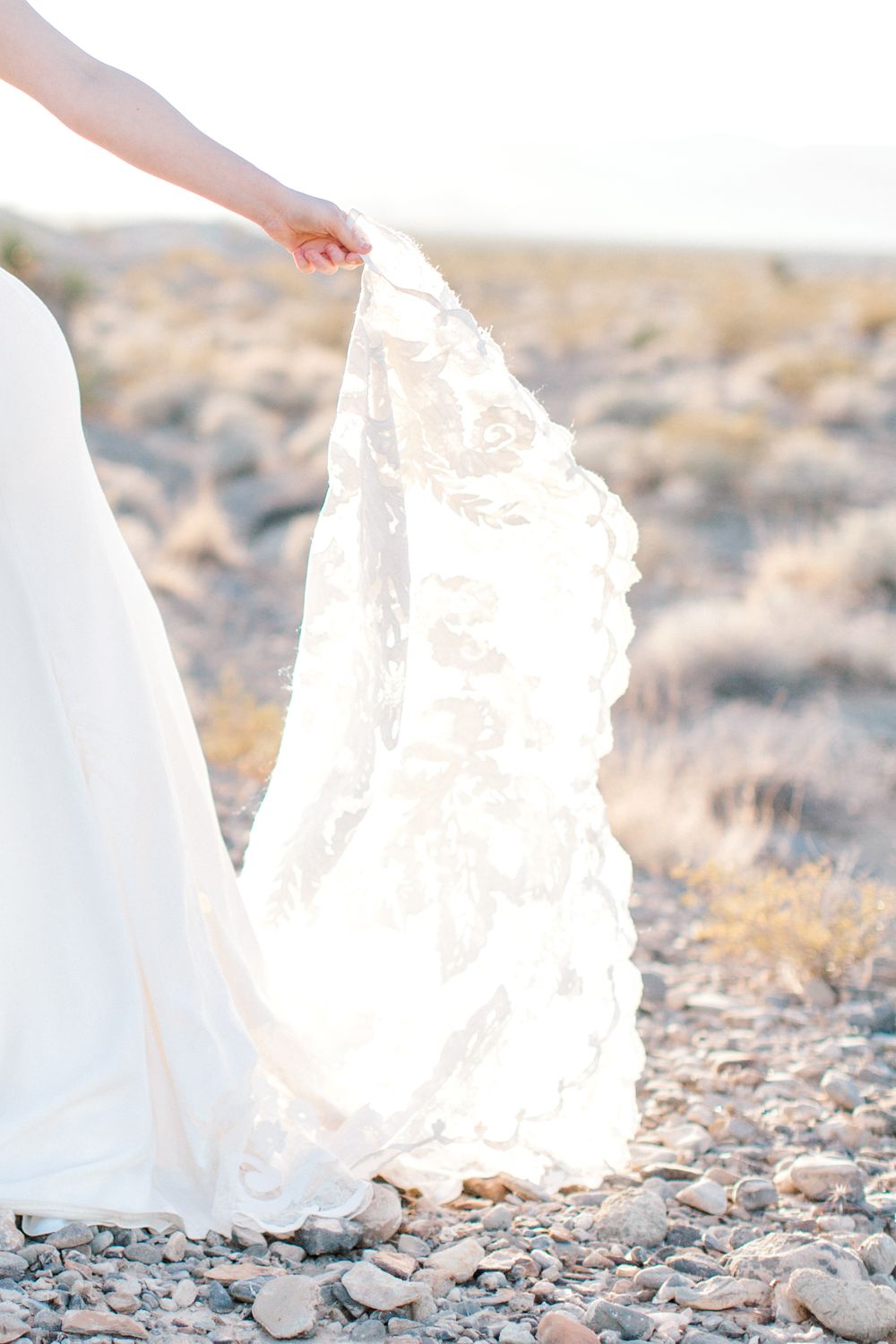 Close-up details of a flowing white wedding dress train against rocky desert terrain.