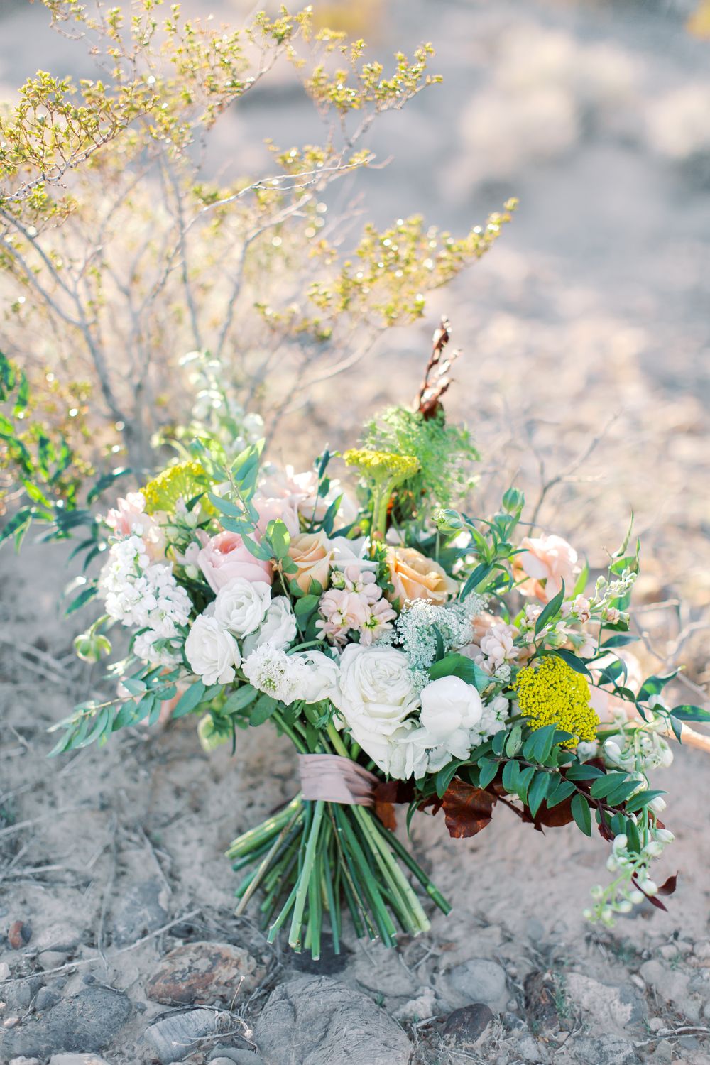 A romantic wedding bouquet with white roses, pink flowers, and greenery arranged on sandy ground with yellow flowers in background.