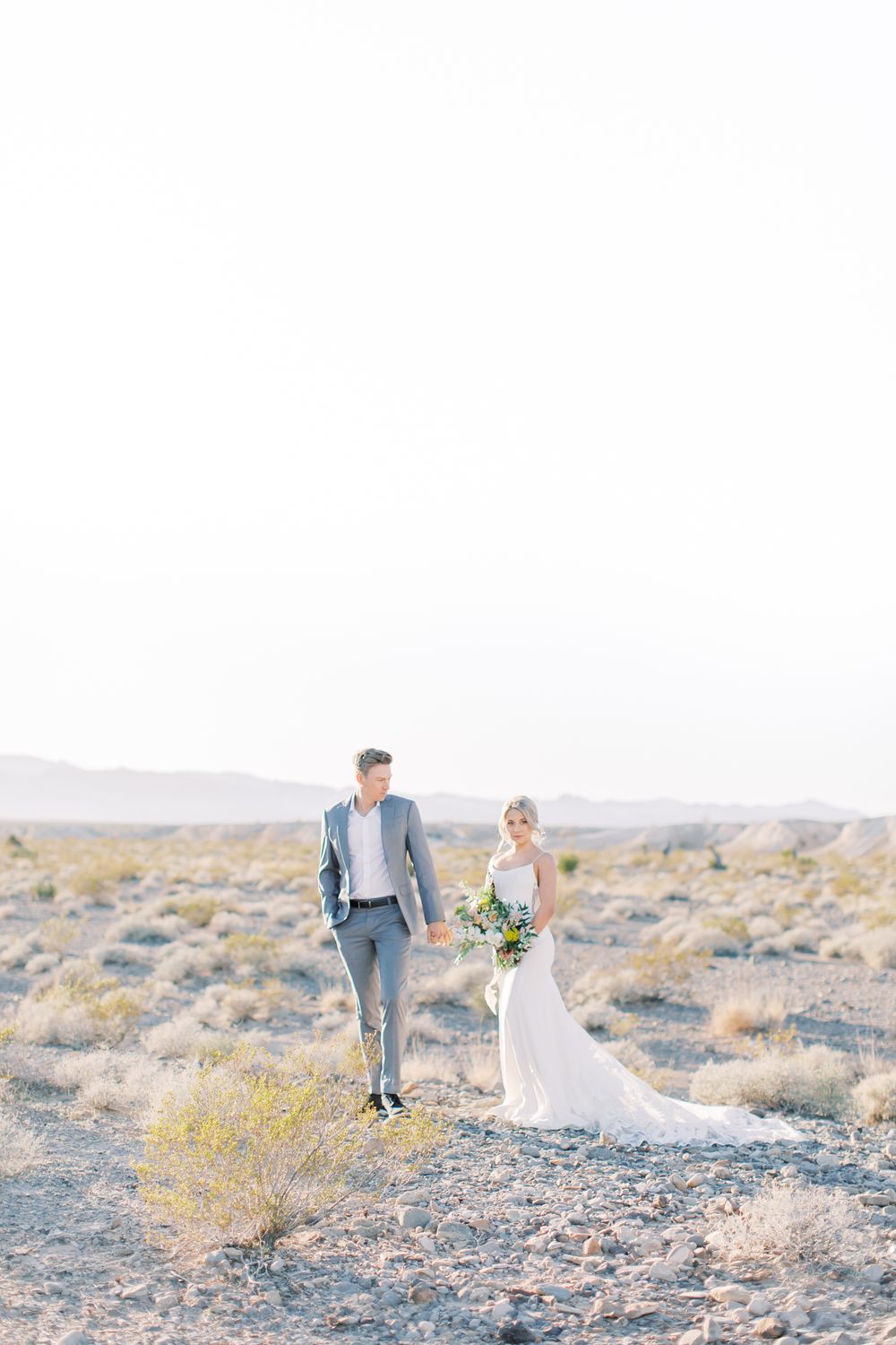 A wedding couple walks together across a desert landscape with yellow wildflowers and mountains in the distance.