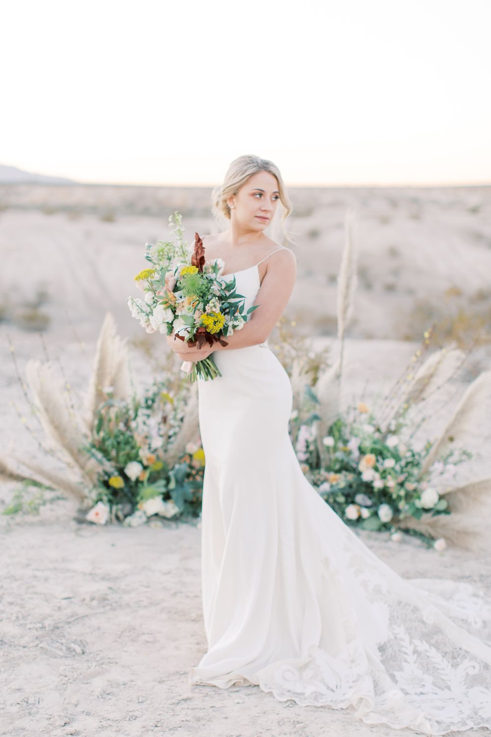 Elegant bride in fitted white gown poses in desert setting with lush bouquet and pampas grass floral arrangement at sunset.