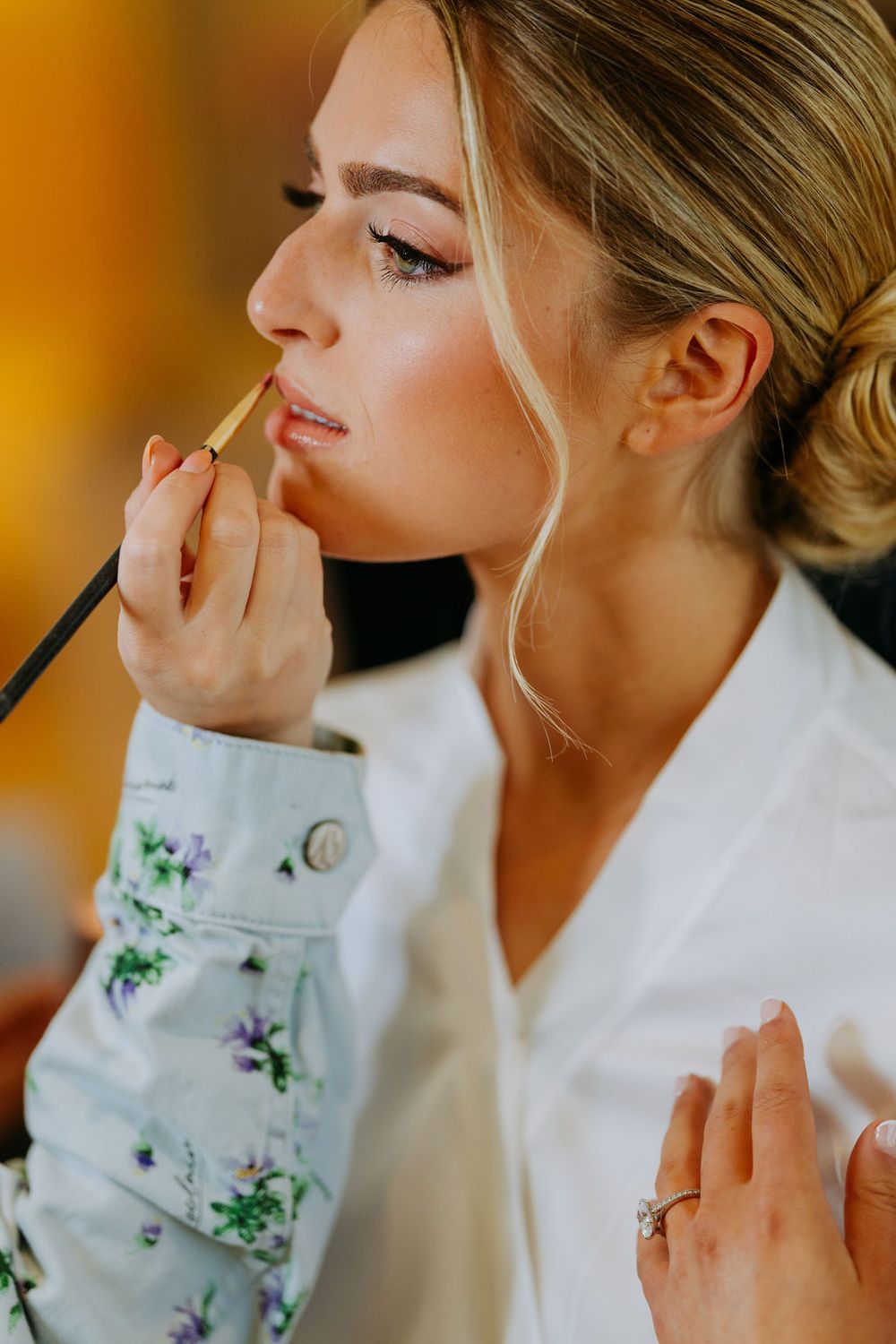 A bride getting her makeup professionally applied during wedding preparations with a floral robe and elegant updo hairstyle.