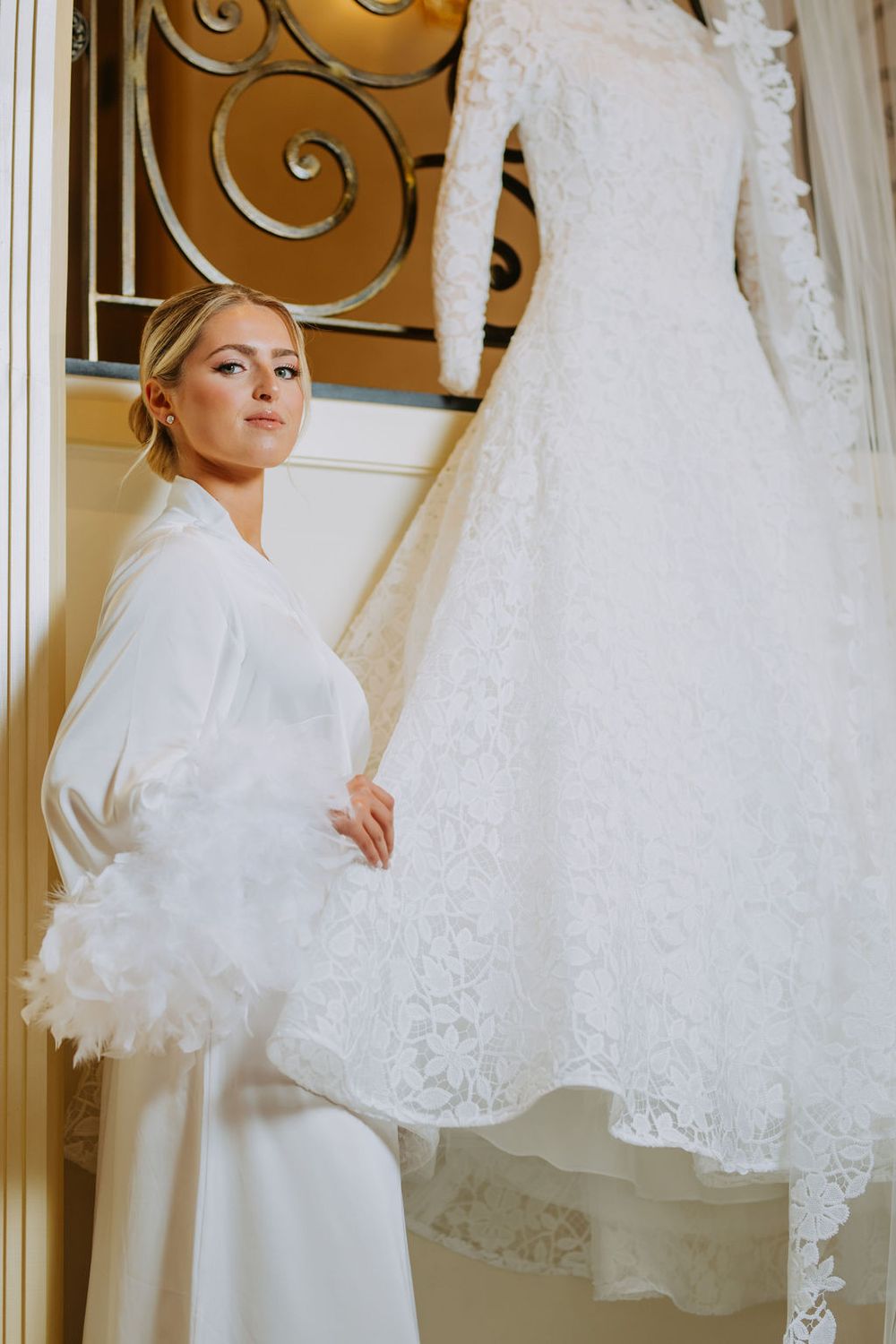 A bride in a white robe examines delicate lace wedding dresses hanging in a bridal boutique.