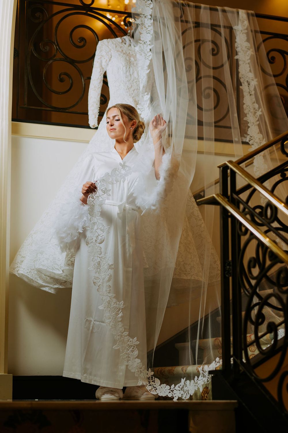 A wedding dress and veil hang elegantly against an ornate staircase with wrought iron details in warm lighting.