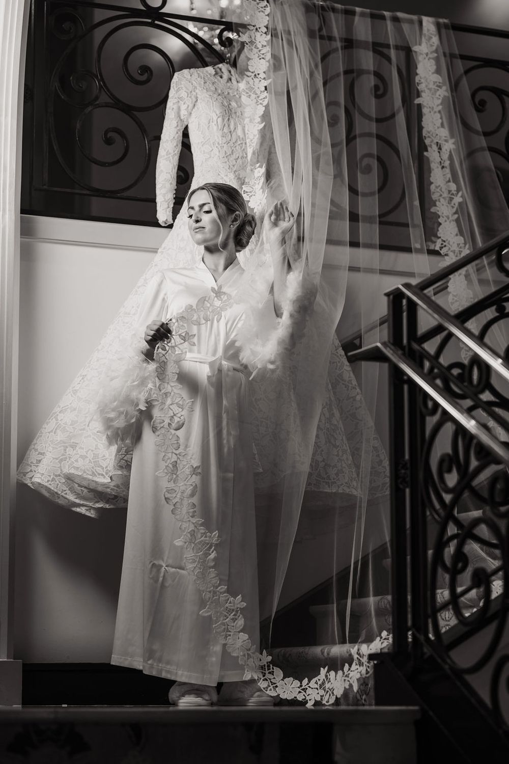 A wedding dress and veil hanging elegantly against a dark staircase with ornate iron railings in a dramatic black and white shot.