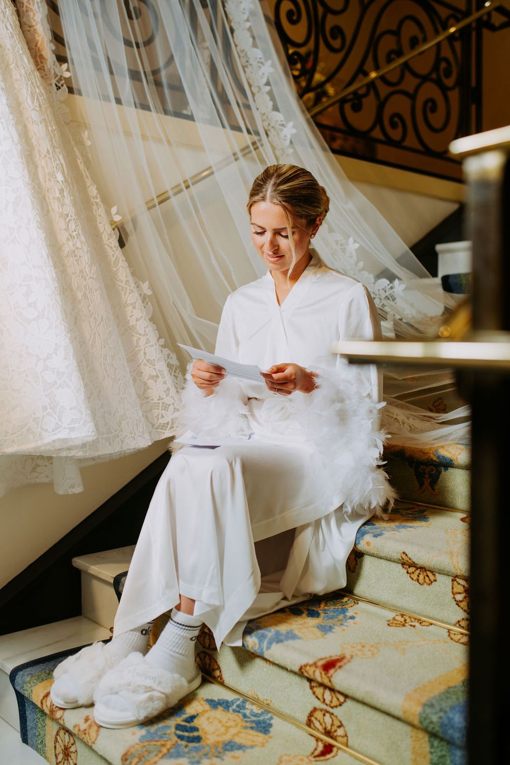 A bride in a white robe sits on a patterned carpet while her wedding dress hangs above in natural light.