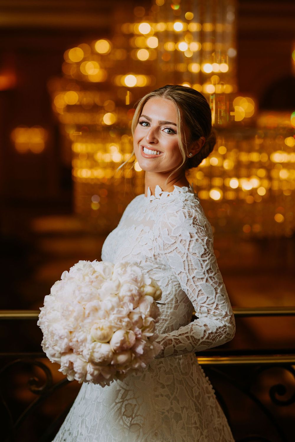 A bride in a lace wedding gown poses with a white floral bouquet against a warm glowing background.