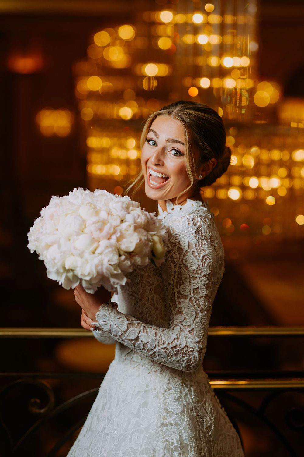 A bride in an elegant white lace dress holds a luxurious bouquet of white flowers against a golden bokeh backdrop.