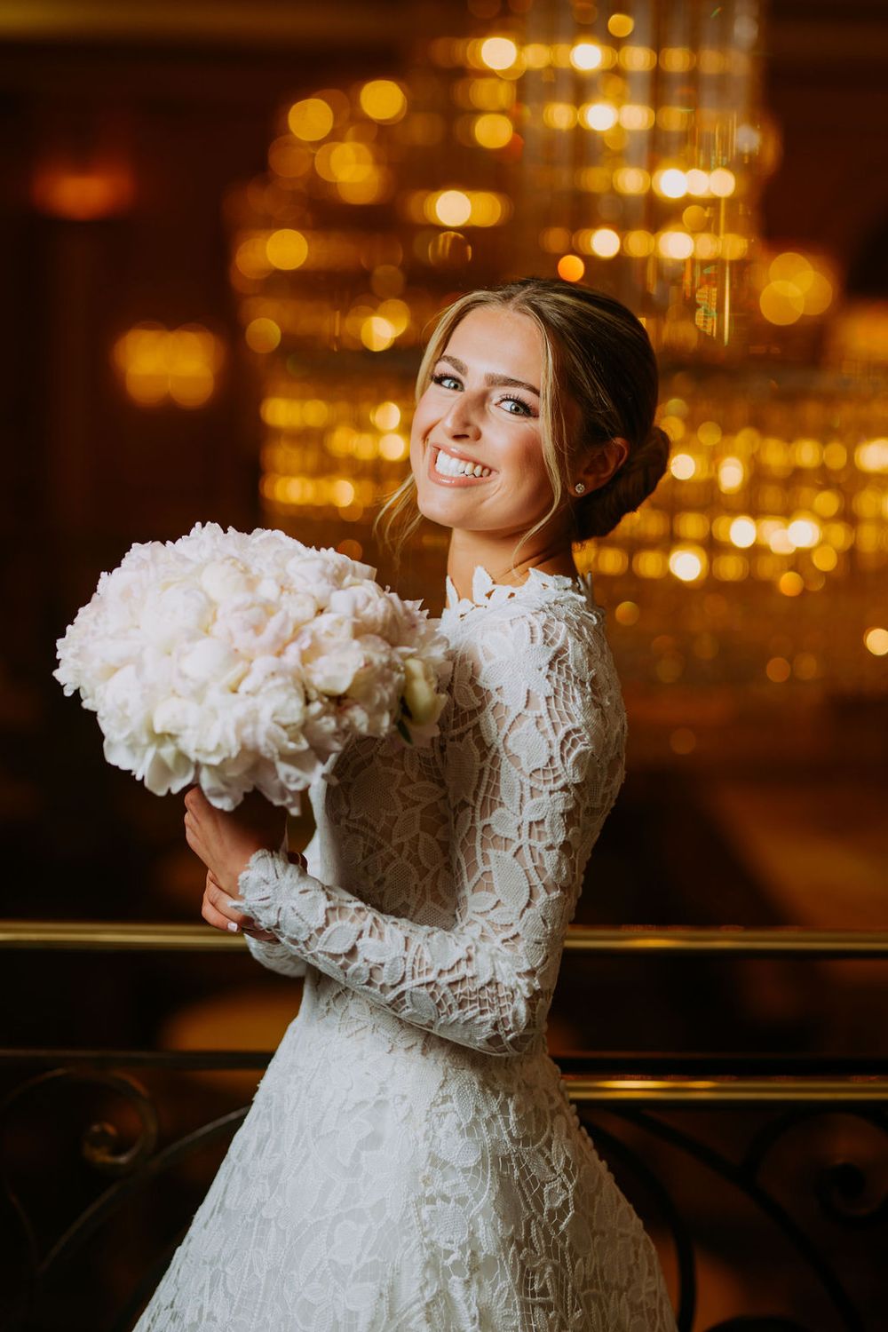A bride in a lace long sleeve wedding dress holds a white bouquet against a bokeh city lights background at night.