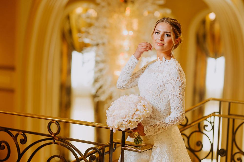 Bride in elegant white lace gown poses in ornate gold and white hallway with wrought iron railing details.