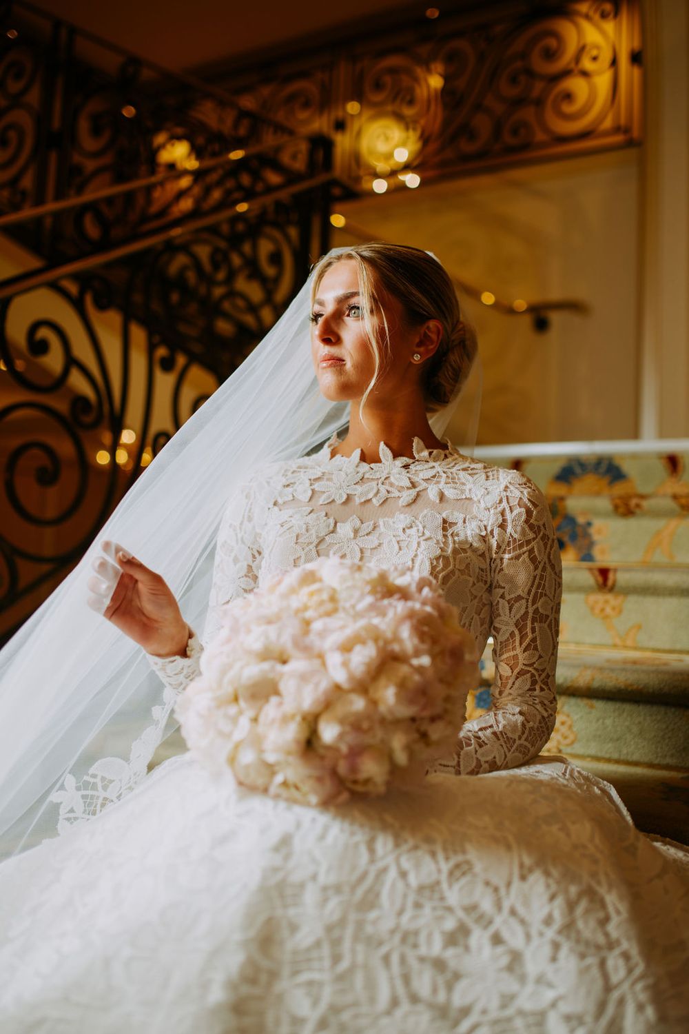 A bride in an elegant lace wedding gown with long sleeves sits on ornate stairs under warm lighting.