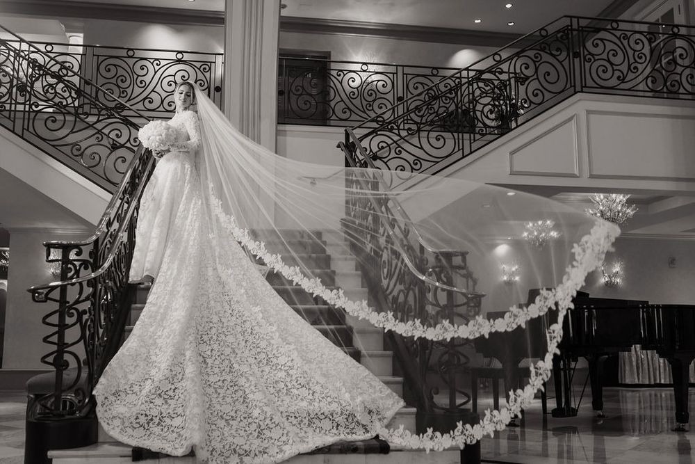A wedding dress with a flowing cathedral veil is displayed in an elegant venue with ornate black iron railings.