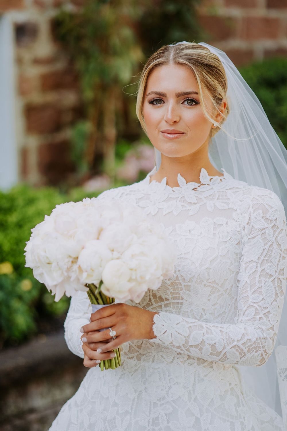 Bride in elegant long-sleeved lace wedding gown holds white rose bouquet against brick wall and greenery.