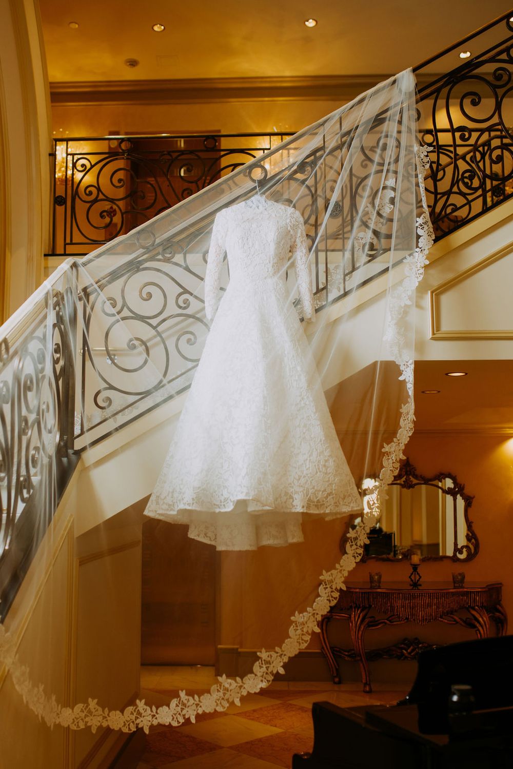 An elegant white lace wedding dress and long veil hang gracefully on an ornate wrought iron staircase railing.