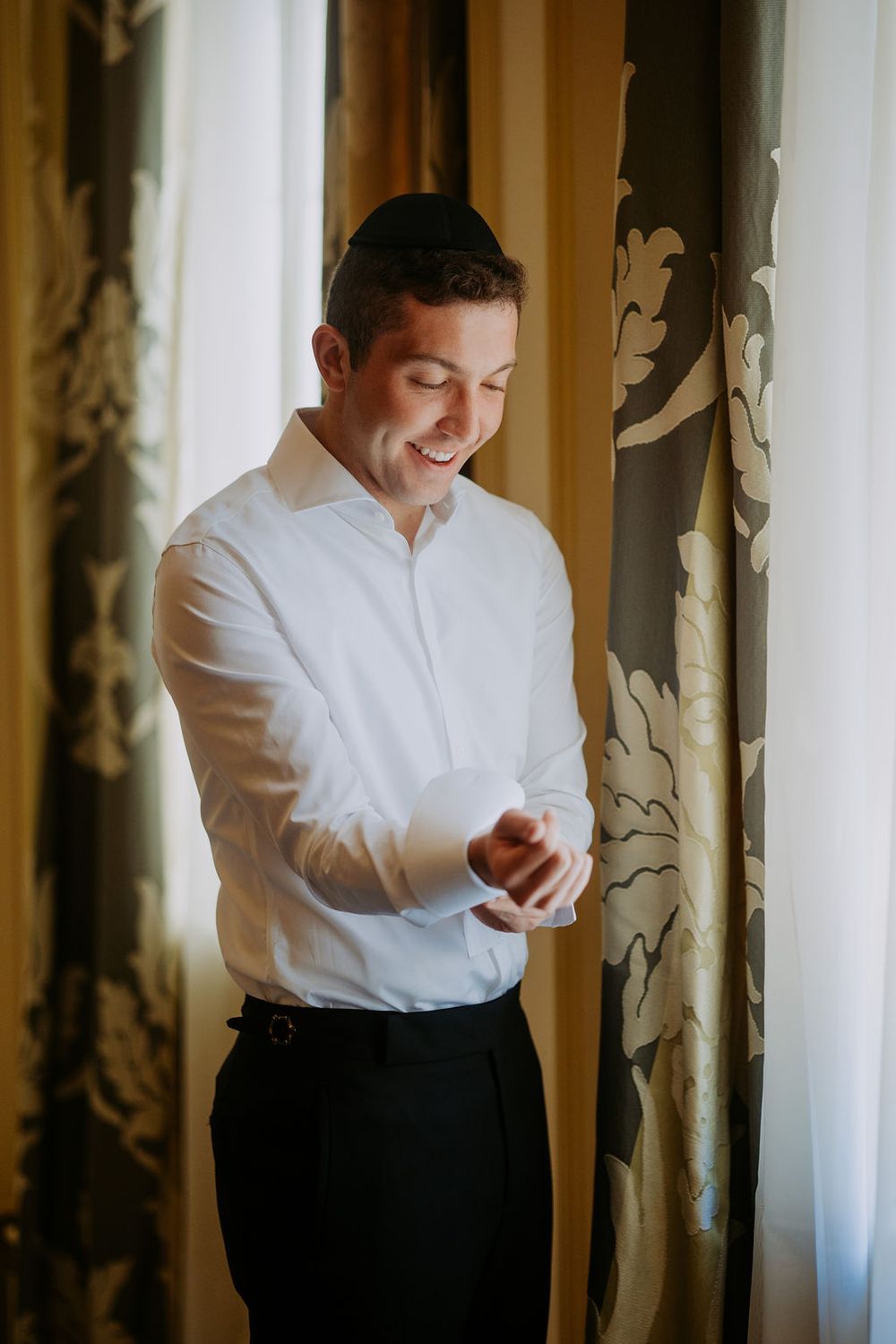 A person in a white dress shirt and black pants smiles while looking down at something in their hands.
