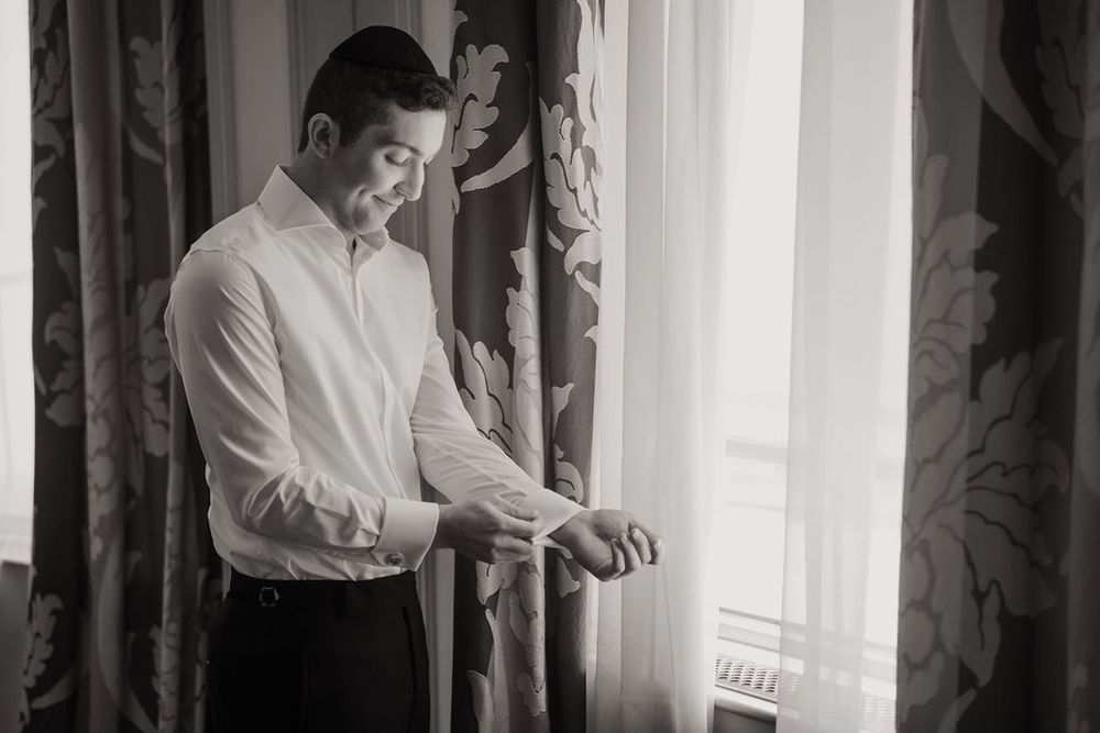 A person in a white shirt stands near a window with sheer curtains, adjusting their sleeve cuff.