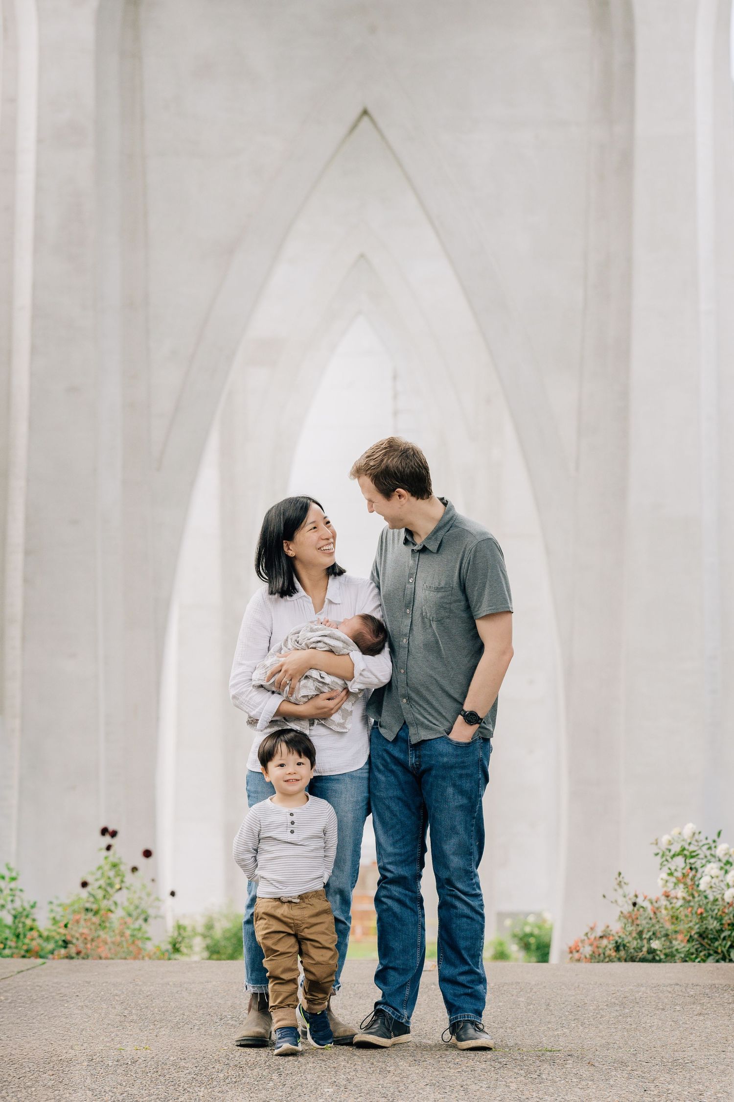 Happy mixed-race family of four posing under white arched bridge with newborn baby and toddler son.