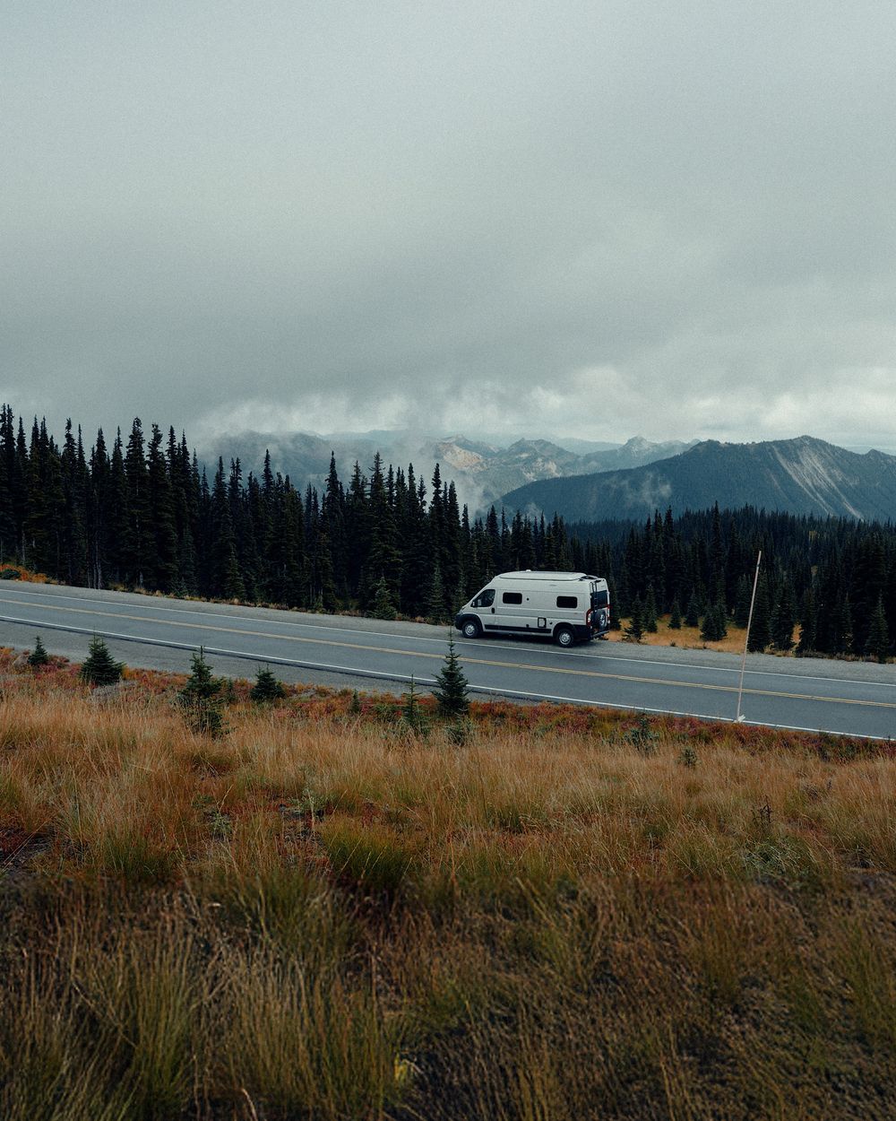 RV camper parked on mountain road with snow-capped peaks and grassland in the background.