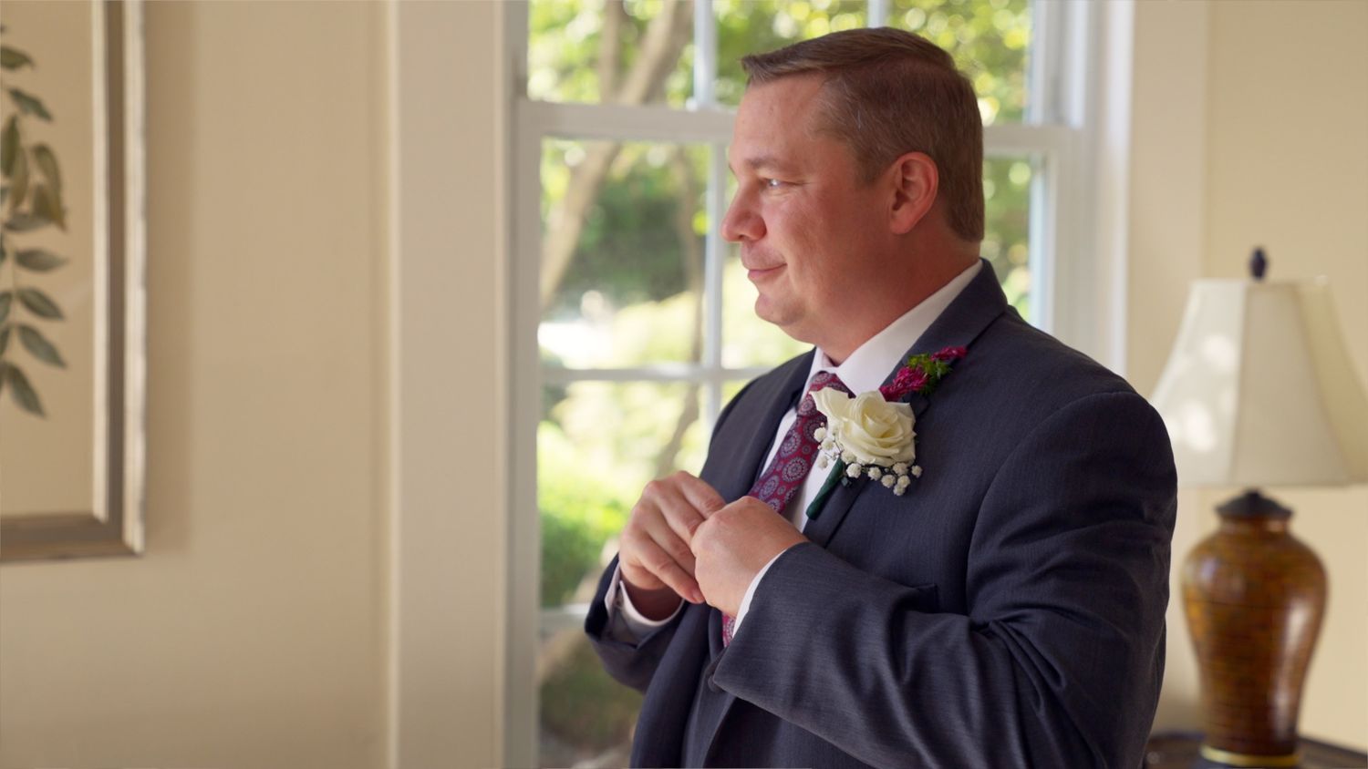A groom in a navy suit adjusts his tie while wearing a white boutonniere before a wedding ceremony.