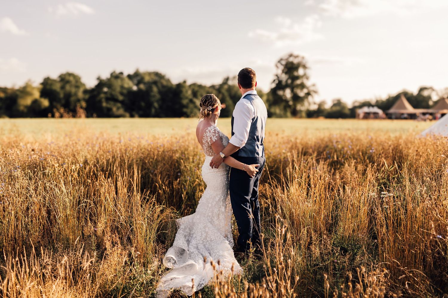 A couple stands together in a golden wheat field at sunset, her white dress flowing in the warm evening light.