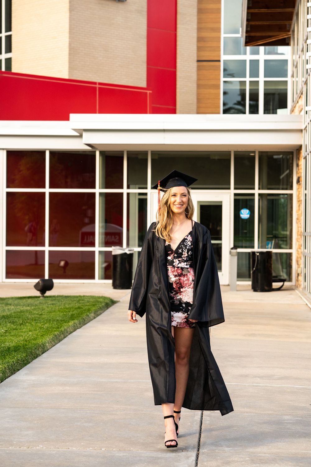 Graduation portrait of a college student in cap and gown with dress showing in front walking in front of her dorm.