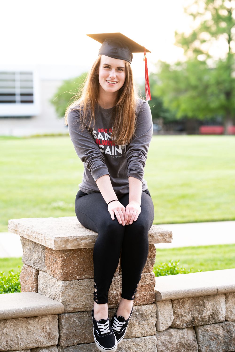 Graduation portrait of a college student in cap sitting on a rock wall in front of a green field.