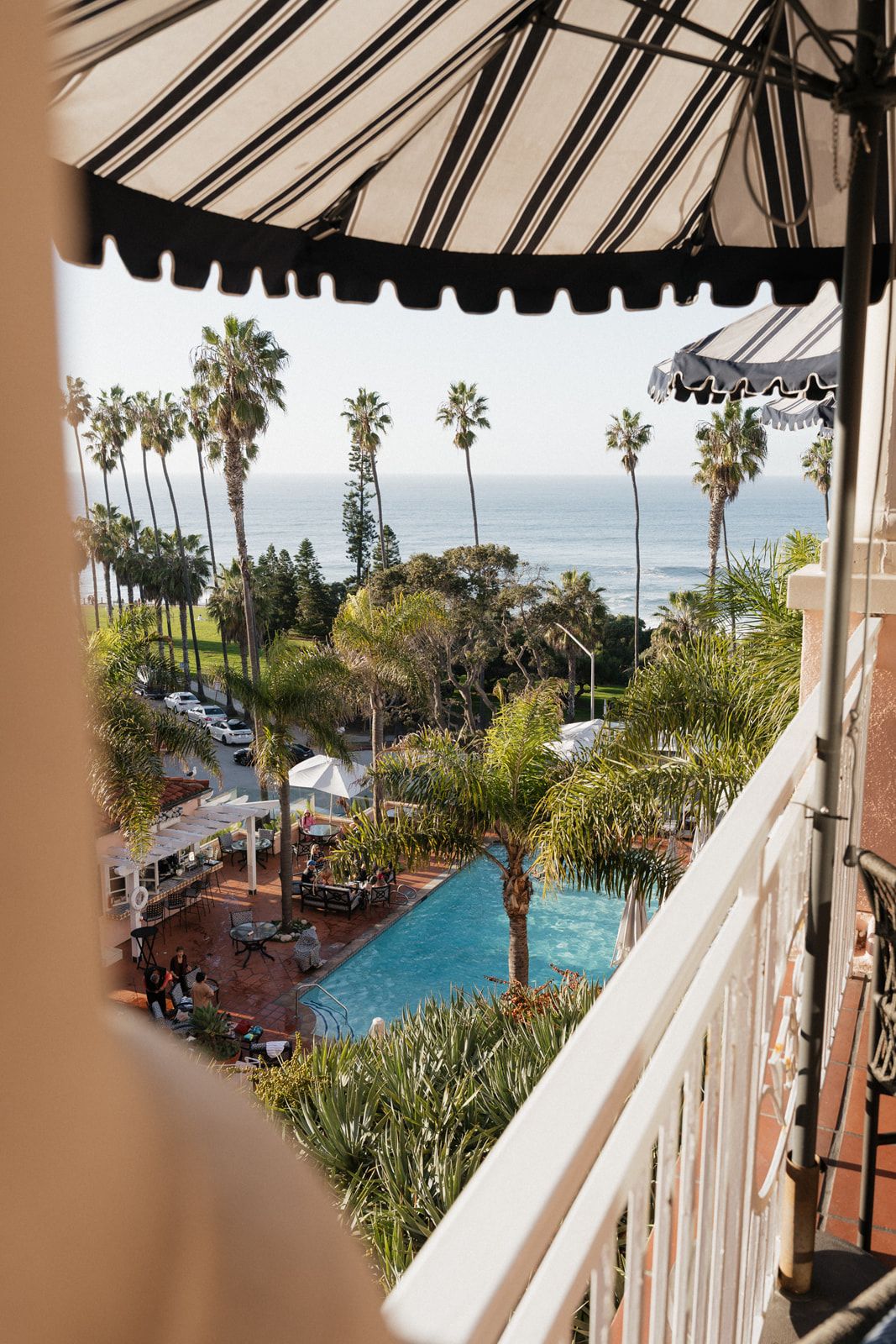 View of a resort pool and palm trees from a balcony with striped awning overlooking the ocean on a sunny day.